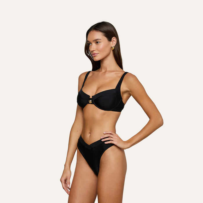 A woman poses against a plain light background, facing left with one hand on her hip, wearing the Adrienne Bikini Top—a black underwire balconette bikini top with thin straps and gold detailing.
