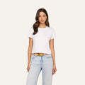 A woman with long brown hair wears the Aileen Cotton Crewneck Tee in plain white, paired with light blue jeans and a tan belt, standing against a plain light background.