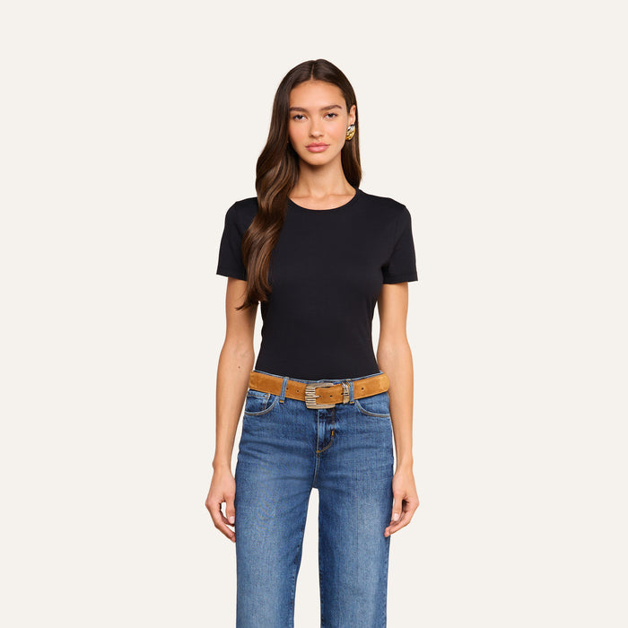 A woman with long brown hair wears the Aileen Cotton Crewneck Tee in black with a relaxed fit, paired with blue jeans and a brown belt, standing against a plain light background.