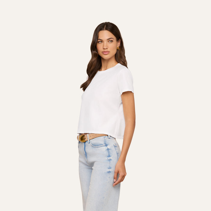 Woman with long brown hair wearing the Aileen Cotton Crewneck Tee, a plain white crewneck top made from soft Supima cotton, paired with light blue jeans. She stands sideways with one hand by her side against a plain light background.