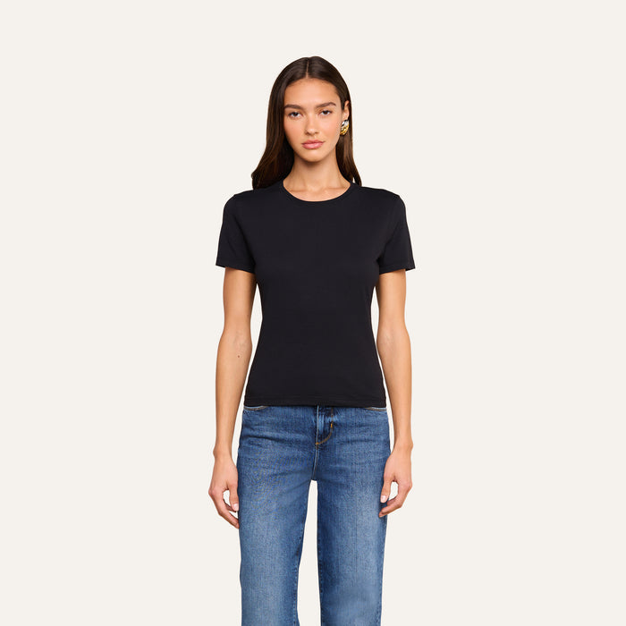A woman with long brown hair is wearing the Aileen Cotton Crewneck Tee in black, made from Supima cotton, paired with blue jeans. She stands facing forward against a plain off-white background.