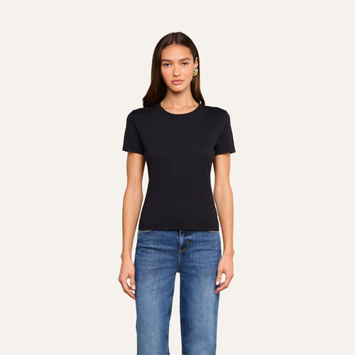A woman with long brown hair is wearing the Aileen Cotton Crewneck Tee in black, made from Supima cotton, paired with blue jeans. She stands facing forward against a plain off-white background.