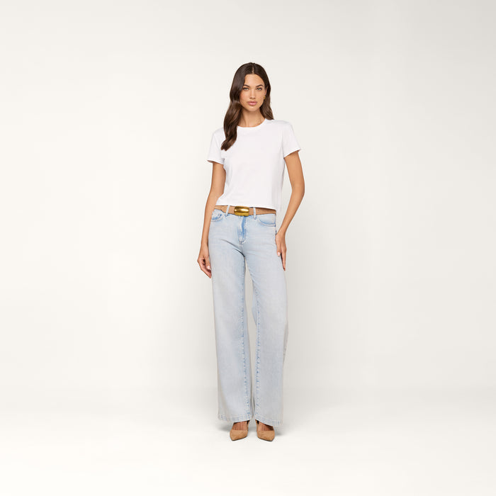 A woman stands against a plain background, wearing the Aileen Cotton Crewneck Tee in white, paired with light blue wide-leg jeans with a brown belt and tan heels. She has long brown hair and a neutral expression.