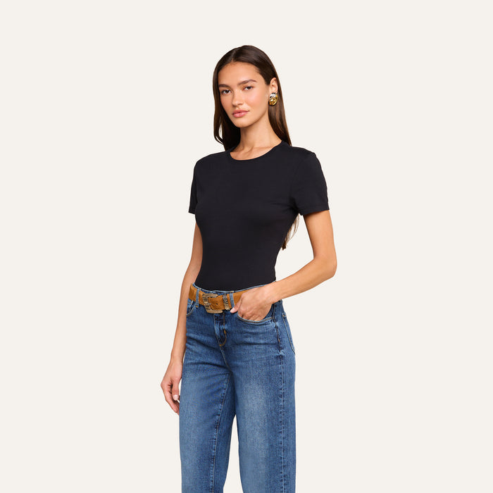 A woman stands against a plain light background, wearing the Aileen Cotton Crewneck Tee in black with blue jeans. She has long dark hair, gold earrings, and one hand in her pocket.
