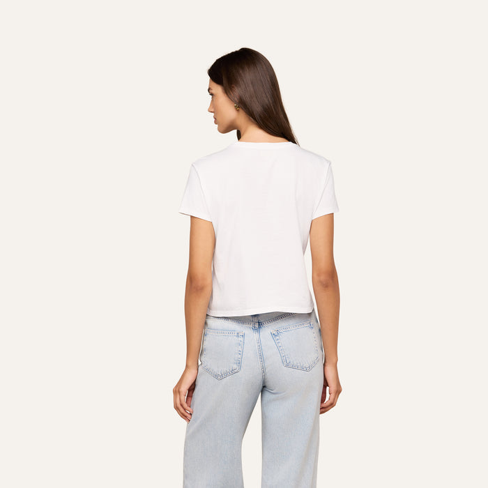 A woman with long brown hair stands with her back to the camera, wearing a white Aileen Cotton Crewneck Tee and light blue jeans against a plain light background.
