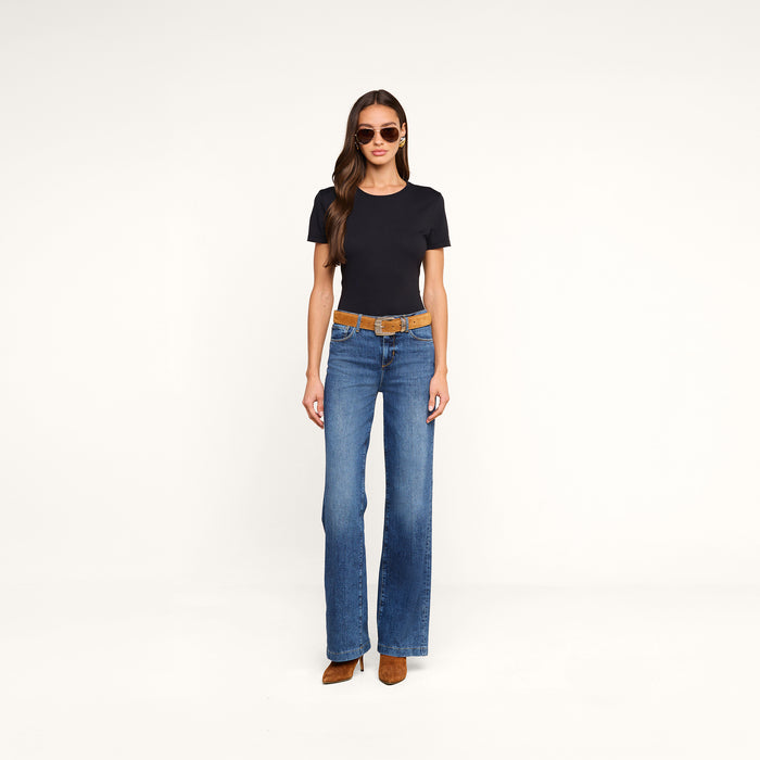 Woman wearing sunglasses, the Aileen Cotton Crewneck Tee in black with a relaxed fit, blue flare jeans with a tan belt, and brown shoes stands against a plain white background.