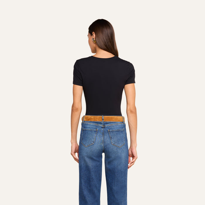 A person with long brown hair, wearing the Aileen Cotton Crewneck Tee in black with a relaxed fit and blue jeans, stands facing away from the camera against a plain light background.