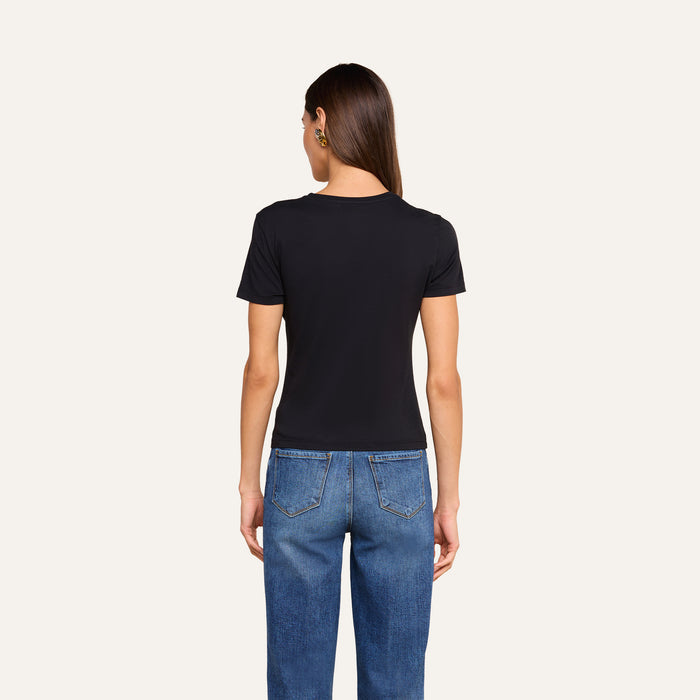 A woman is standing with her back to the camera, wearing the Aileen Cotton Crewneck Tee in black paired with blue jeans. Her straight brown hair is worn down, and her arms are relaxed at her sides. The background is plain and light-colored.
