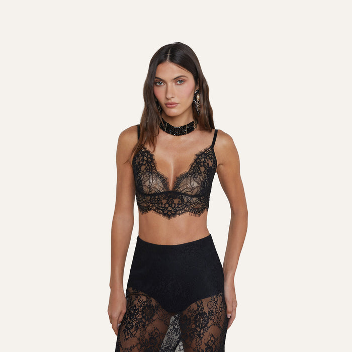 A woman wears the Aislin Lace Bralette, featuring adjustable straps and scalloped edges, paired with a black lace skirt in sheer stretch lace, as she poses against a plain light background.