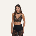 A woman wears the Aislin Lace Bralette, featuring adjustable straps and scalloped edges, paired with a black lace skirt in sheer stretch lace, as she poses against a plain light background.