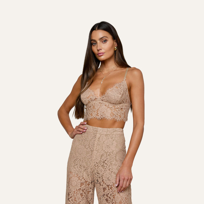 A woman wearing the Aislin Lace Bralette with adjustable straps and high-waisted lace pants featuring scalloped edges stands against a plain light background, looking at the camera with a relaxed expression.