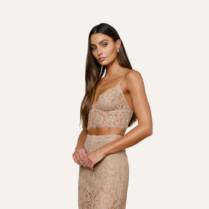 A woman with long brown hair wears the Aislin Lace Bralette, a nude-colored lace crop top featuring sheer stretch lace and delicate scalloped edges. She poses with hands clasped, her expression neutral, against an off-white background.