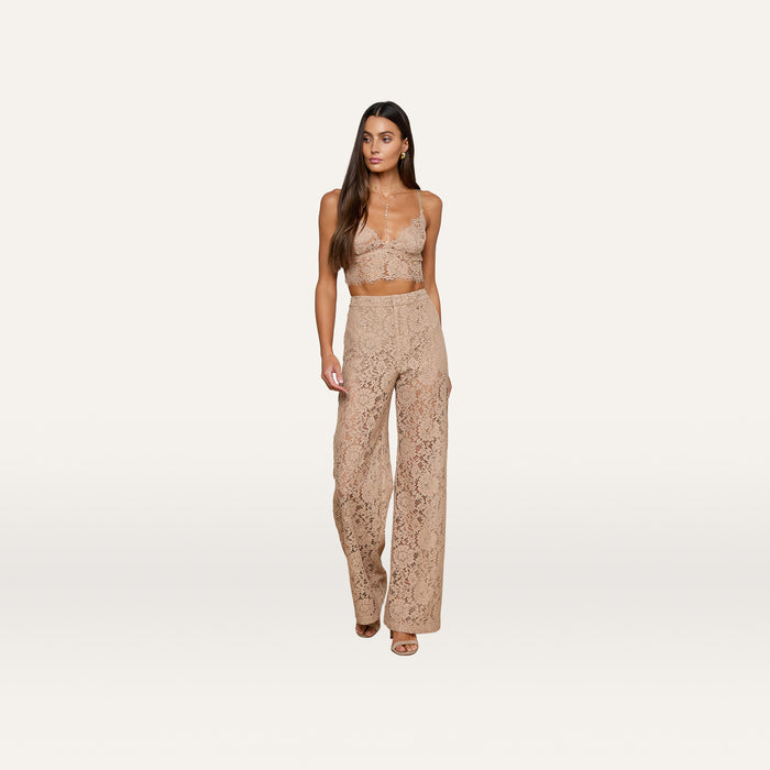A woman wearing the Aislin Lace Bralette in beige with adjustable straps and matching high-waisted lace pants, both featuring sheer stretch lace and scalloped edges, stands against a plain white background.