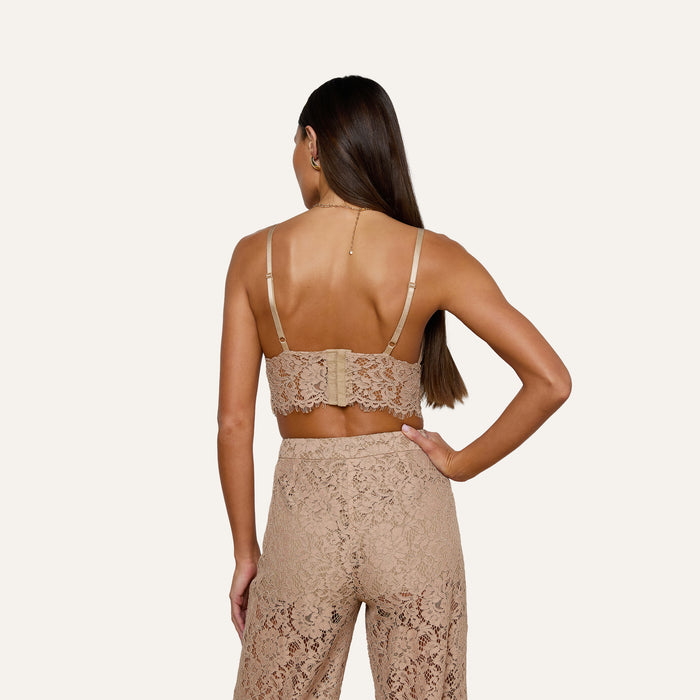 A woman with long brown hair stands with her back to the camera against a plain light background, wearing the Aislin Lace Bralette in beige—a sheer stretch lace bralette with adjustable straps—paired with matching lace pants.