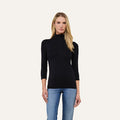 A woman with long blonde hair wears the Aja Turtleneck, a slim-fit black top with three-quarter sleeves, paired with blue jeans and standing against a plain light background.