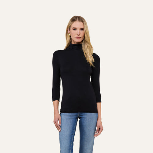 A woman with long blonde hair wears the Aja Turtleneck, a slim-fit black top with three-quarter sleeves, paired with blue jeans and standing against a plain light background.