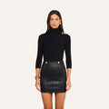 A woman stands facing forward, wearing the Aja Turtleneck in luxe black modal with gold buttons on the shoulder, paired with a black leather mini skirt featuring gold button accents. Her long brown hair is complemented by gold hoop earrings and bracelets, creating a sophisticated look ideal for transitioning from fall to winter.