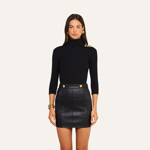 A woman stands facing forward, wearing the Aja Turtleneck in luxe black modal with gold buttons on the shoulder, paired with a black leather mini skirt featuring gold button accents. Her long brown hair is complemented by gold hoop earrings and bracelets, creating a sophisticated look ideal for transitioning from fall to winter.