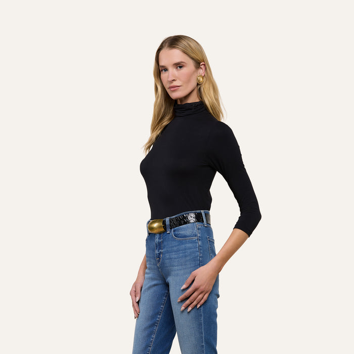 A woman stands against a plain light background, facing slightly to the side with one hand on her hip, wearing the slim-fit Aja Turtleneck in black—crafted from microfiber fabrication—paired with blue jeans and a black belt.
