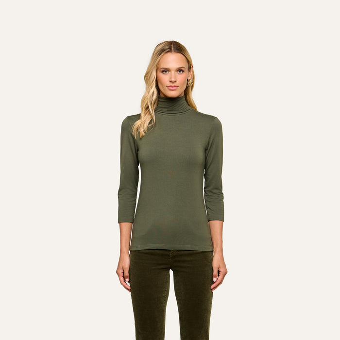 A woman wearing the Aja Turtleneck—a slim fit olive green top with three-quarter sleeves—along with matching pants stands facing forward against a plain light background.