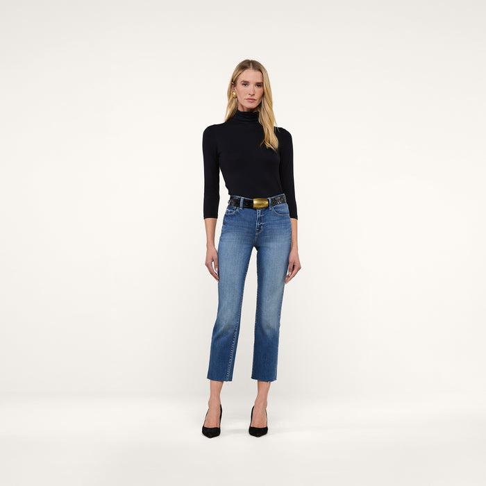 A woman stands against a plain white background, wearing the slim-fit black Aja Turtleneck, blue high-waisted jeans with a black belt, and black high heels. She faces forward with a neutral expression and her hands by her sides.