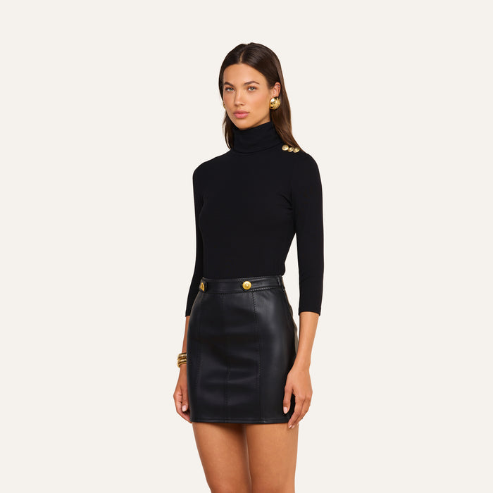 Wearing the Aja Turtleneck—a luxe modal black top featuring gold buttons on the shoulder—paired with a black leather mini skirt adorned with gold buttons, this look creates a chic statement against a plain light background. Perfect for refreshing your fall-to-winter wardrobe.