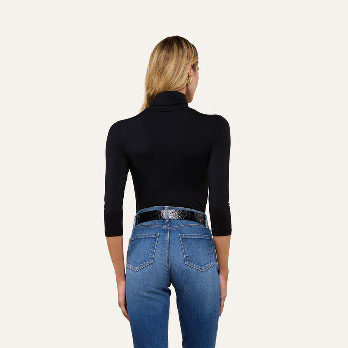 A woman with long blonde hair, wearing the Aja Turtleneck in black along with blue jeans and a black belt, stands with her back to the camera against a plain background.