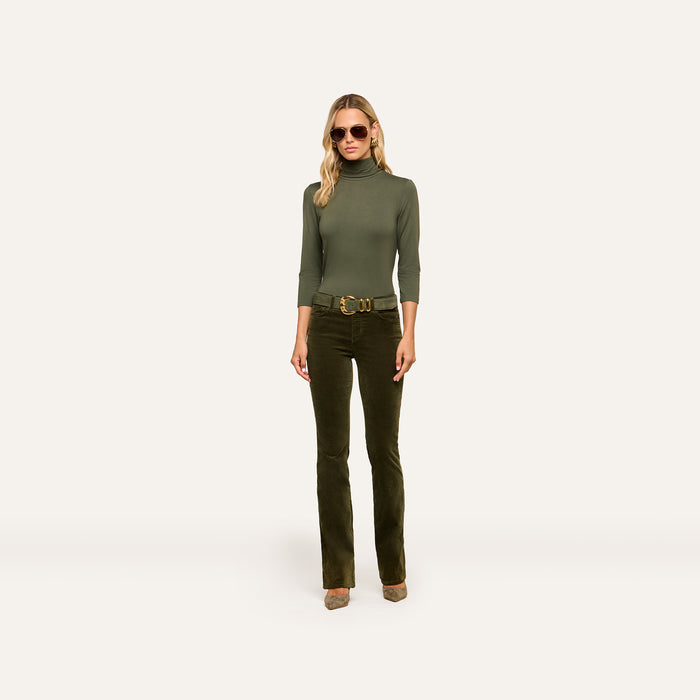 A woman wearing sunglasses, the Aja Turtleneck in a slim fit green style, dark green flared pants with a belt, and heels stands against a plain light background.
