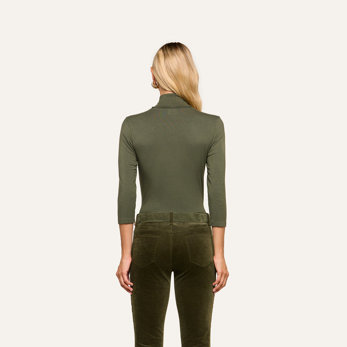 A woman with long blonde hair stands facing away, wearing the Aja Turtleneck in olive green with three-quarter sleeves and slim fit, paired with dark olive velvet pants, against a plain light background.