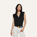 Woman wearing the Akari Bodysuit—a sleeveless black top with subtle shoulder pads—paired with white pants, a black belt with a gold buckle, gold necklace, and earrings, standing against an off-white background.