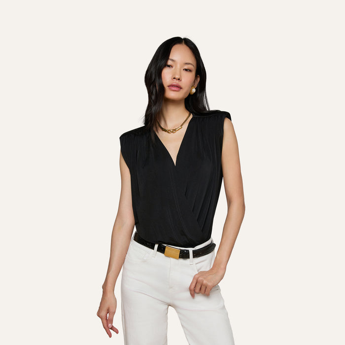 Woman wearing the Akari Bodysuit—a sleeveless black top with subtle shoulder pads—paired with white pants, a black belt with a gold buckle, gold necklace, and earrings, standing against an off-white background.