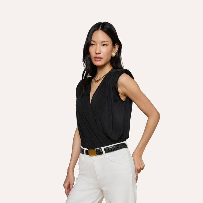 A woman with long black hair wears the Akari Bodysuit—a sleeveless black liquid jersey top with a deep V-neck—paired with white pants. She accessorizes her look with gold earrings, a necklace, and a black belt featuring a gold buckle while posing against a plain light background.