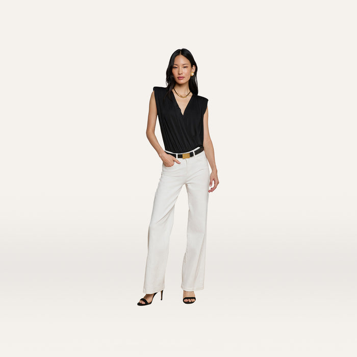 A woman stands against a plain background in a sleeveless black Akari Bodysuit, paired with white wide-leg pants, a black belt, black heels, and a gold necklace. With one hand in her pocket, she poses in a relaxed manner.