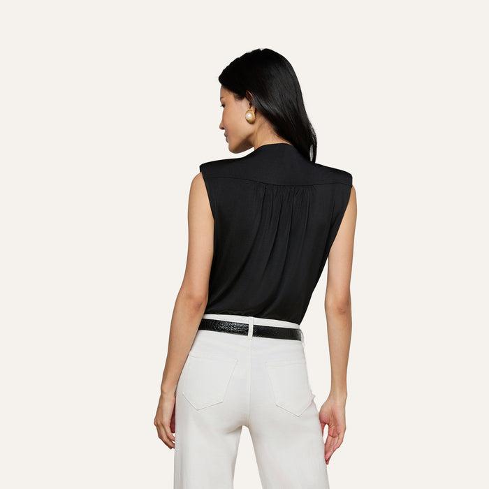 A woman stands with her back to the camera, wearing the sleeveless black Akari Bodysuit, paired with white pants, a black belt, and large round earrings against a plain light background.