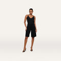 A woman stands confidently against a plain white background, wearing a black sleeveless top, the Akasha Bermuda Short in high-rise black stretch ponte, and black heeled sandals.
