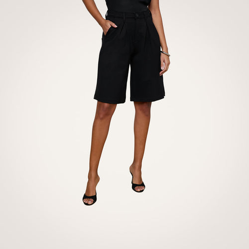 A person wearing the Akasha Bermuda Short—black high rise stretch ponte bermuda shorts—with a black top and black open-toe heels stands against a plain white background. Their hands are in their pockets, and only the lower half of their body is visible.