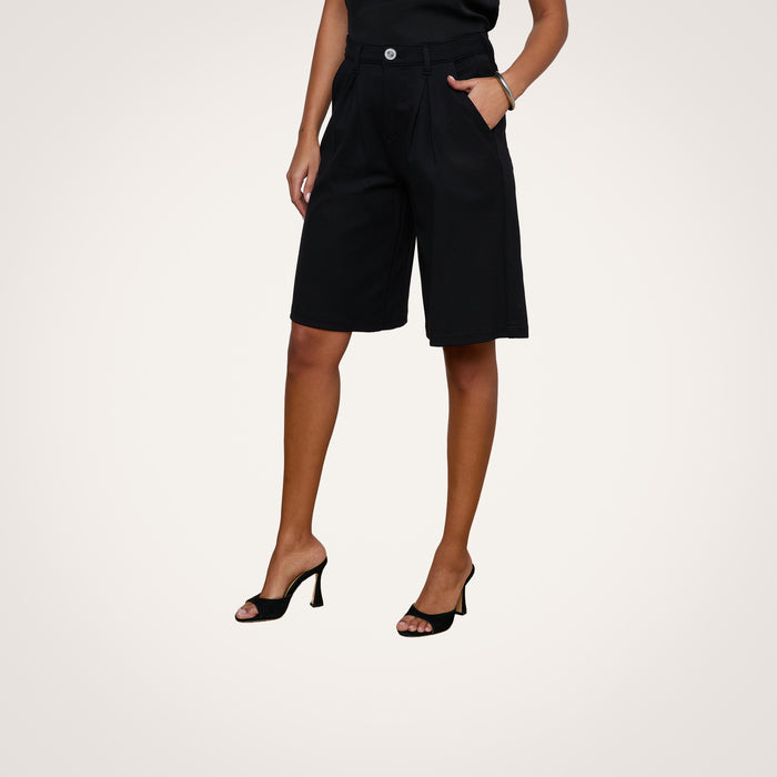A person wearing the Akasha Bermuda Short in black, paired with a black sleeveless top and black heeled sandals, stands with hands in pockets against a plain light background.