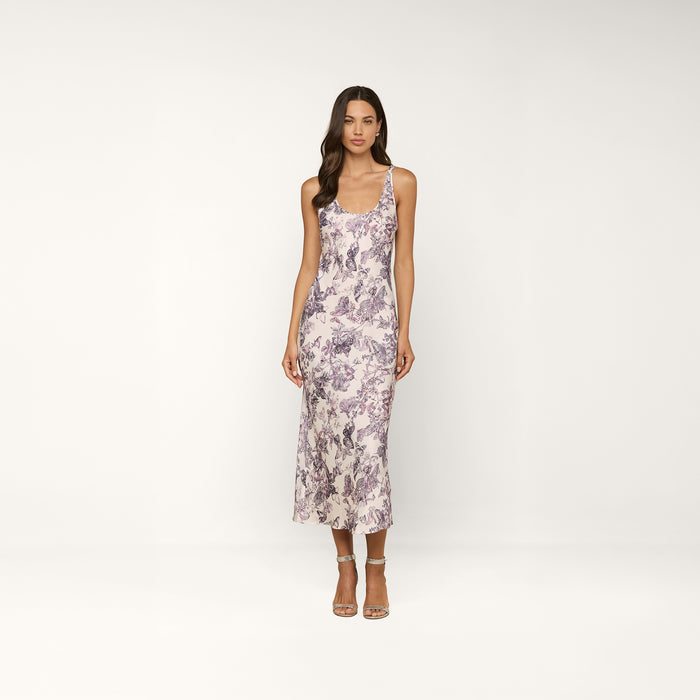 A woman stands in a studio against a plain white background, wearing the fitted, sleeveless Akiya Satin Maxi Dress in white with a purple floral pattern, paired with strappy heeled sandals.