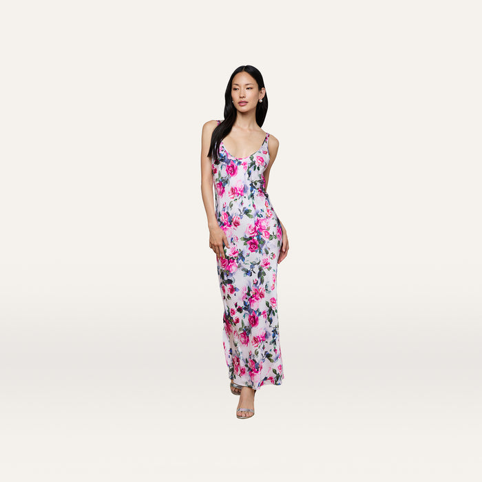 A woman wearing the Akiya Satin Maxi Dress—a sleeveless, white, floor-length bias-cut gown adorned with a pink and blue floral pattern—stands against a plain off-white background.