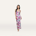 A woman wearing the Akiya Satin Maxi Dress—a sleeveless, white, floor-length bias-cut gown adorned with a pink and blue floral pattern—stands against a plain off-white background.