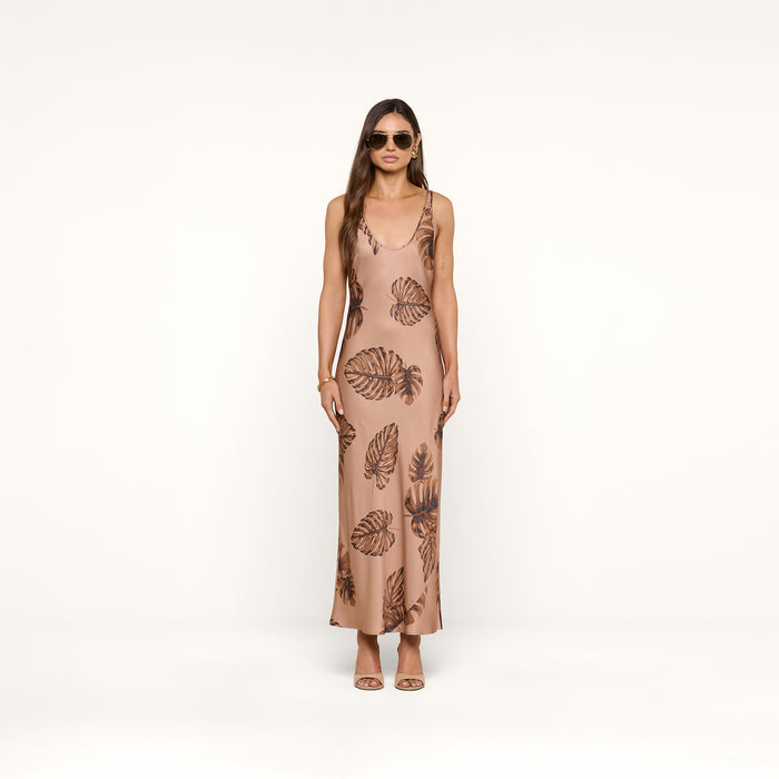 A woman wearing sunglasses and a long, sleeveless tan Akiya Satin Maxi Dress with a brown palm leaf print stands facing forward against a plain white background.