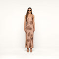 A woman wearing sunglasses and a long, sleeveless tan Akiya Satin Maxi Dress with a brown palm leaf print stands facing forward against a plain white background.