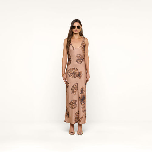 A woman wearing sunglasses and a long, sleeveless tan Akiya Satin Maxi Dress with a brown palm leaf print stands facing forward against a plain white background.