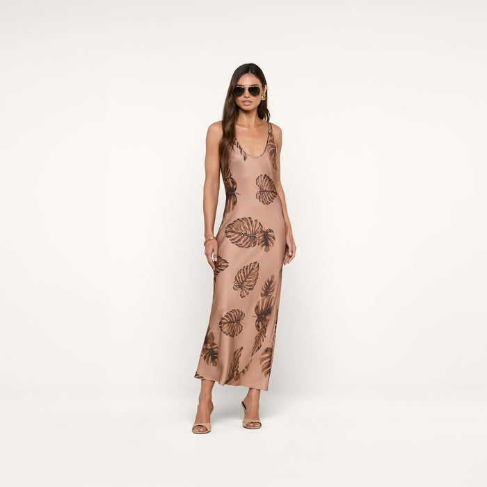 Woman wearing sunglasses and a sleeveless, ankle-length beige Akiya Satin Maxi Dress with a brown palm leaf print, standing against a plain white background. She has long hair and is wearing heeled sandals.