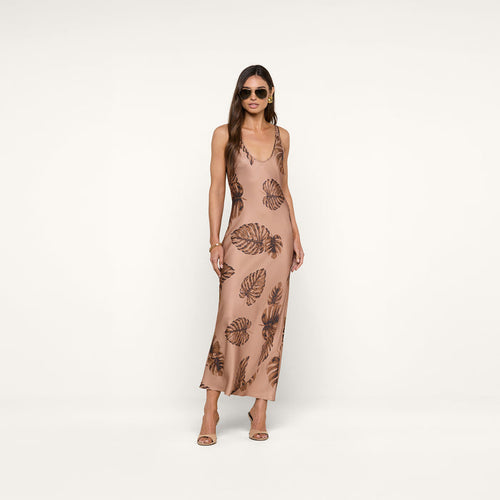 Woman wearing sunglasses and a sleeveless, ankle-length beige Akiya Satin Maxi Dress with a brown palm leaf print, standing against a plain white background. She has long hair and is wearing heeled sandals.