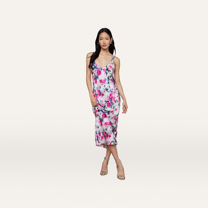Wearing the Akiya Satin Maxi Dress, which features a bias-cut silhouette and pink and purple floral prints, the woman stands against a plain light background in strappy heels.