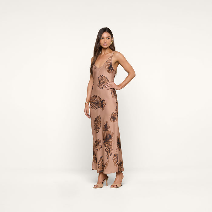 A woman stands wearing the Akiya Satin Maxi Dress, a sleeveless, ankle-length design in brown palm leaf print crafted from bias-cut satin. She has long hair and is styled with brown open-toe heels, posing against a plain white background.
