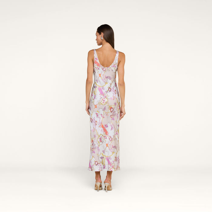 A woman stands against a white background, facing away. She is wearing the Akiya Satin Maxi Dress—an elegant, sleeveless long dress in pastel floral tones—and heeled sandals. Her straight hair falls down her back, and she maintains a neutral posture with her arms relaxed by her sides.