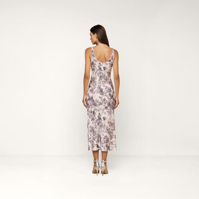 A woman stands with her back to the camera, wearing the sleeveless, ankle-length Akiya Satin Maxi Dress featuring a delicate butterfly print, paired with white heeled sandals. The background is plain and light-colored.