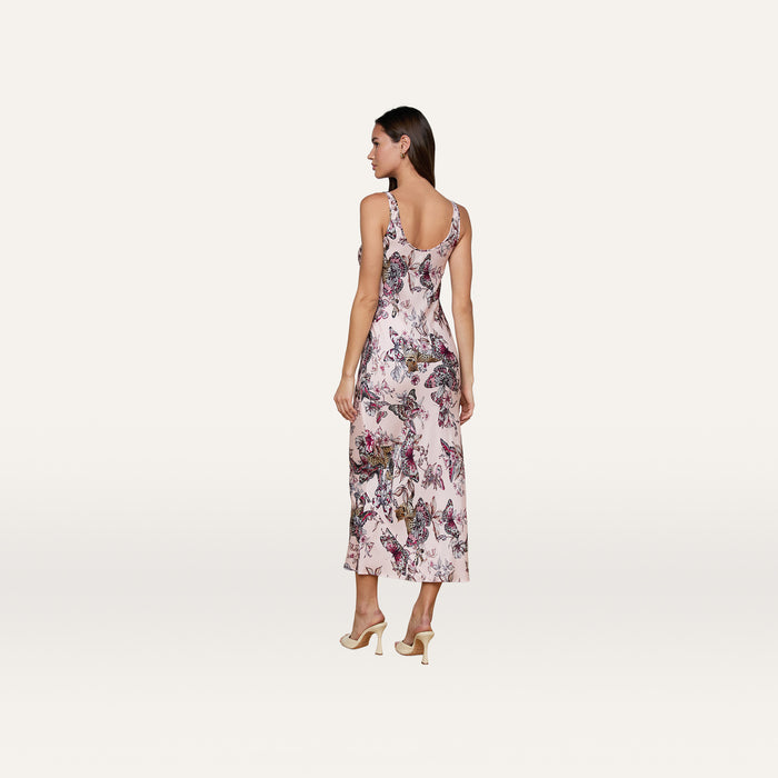 A woman stands facing away against a plain, light background, wearing the Akiya Satin Maxi Dress—a bias-cut satin gown featuring a butterfly jungle print—paired with light-colored high heels.